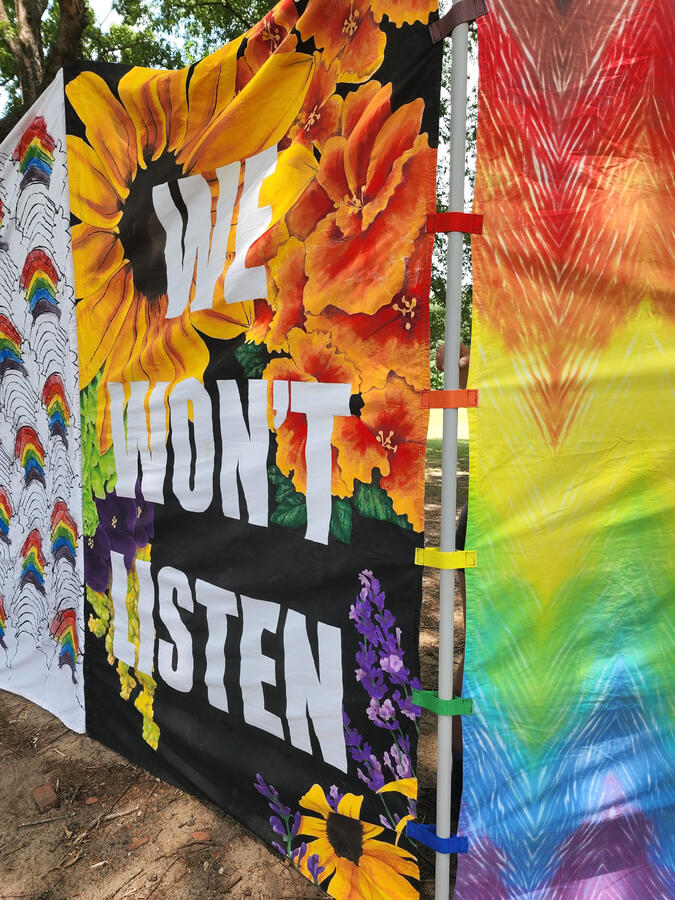 Close up three 8'x5' vertical banners, the far one is white with a screenprinted rainbows pattern, the middle says "WE WON'T LISTEN" on a floral backgrouynd, and the nearest is a zigzag tie dye of the Philly Pride flag